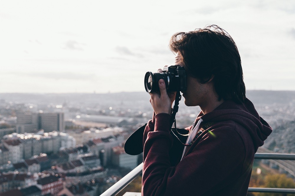 A man is holding up a camera, aiming to take a picture of something out of frame. He's wearing a dark red hoodie and seems to be standing on a balcony or ledge of a very tall building