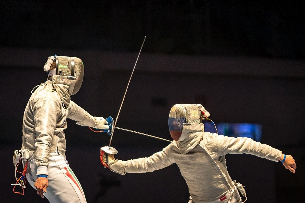 Two people are fencing. Both are wearing proper fencing outfits and facing off against each other in a heated battle