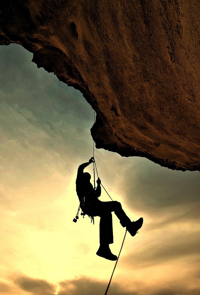 A man in rock climbing gear hangs off the side of a cliff. The sun is sitting in the background creating a silhouette effect on the climber