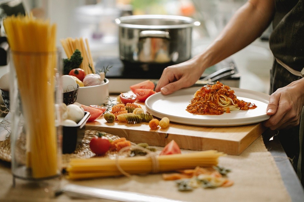 A table has various pastas, tomatoes, and cooking utensils on it. Someone just out of frame is plating some spaghetti onto a plate. Their hand is holding the plate about to lift it up and serve it to someone