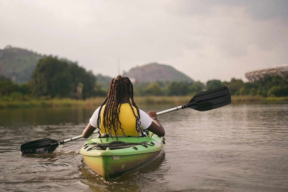 A man with long dreadlocks is sitting in a kayak in an open body of water. He is paddling out into the water