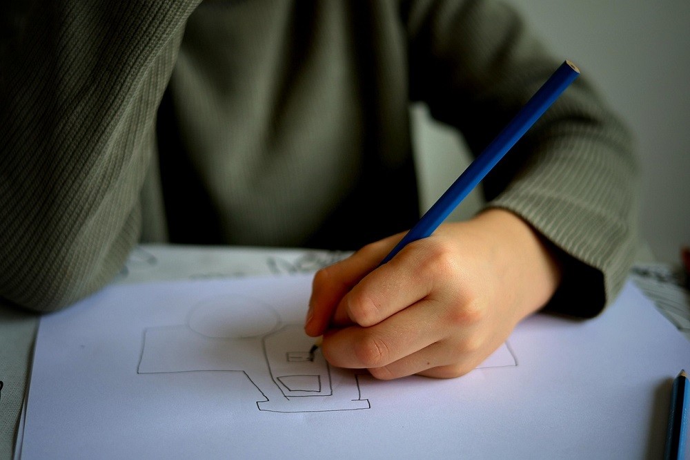 A younger person is drawing a car on a piece of paper. The drawing is very childish and the lines aren't straight, like it's their first time drawing one