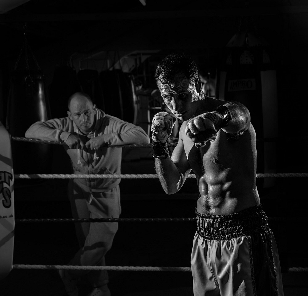 A young male boxer is in the ring. His coach is leaning on the ropes outside of the ring. The coach is focused on the boxer