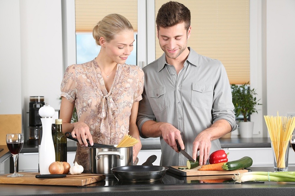 A young couple are in the kitchen preparing a meal together. There are various vegetables on the table along with several pots and pans