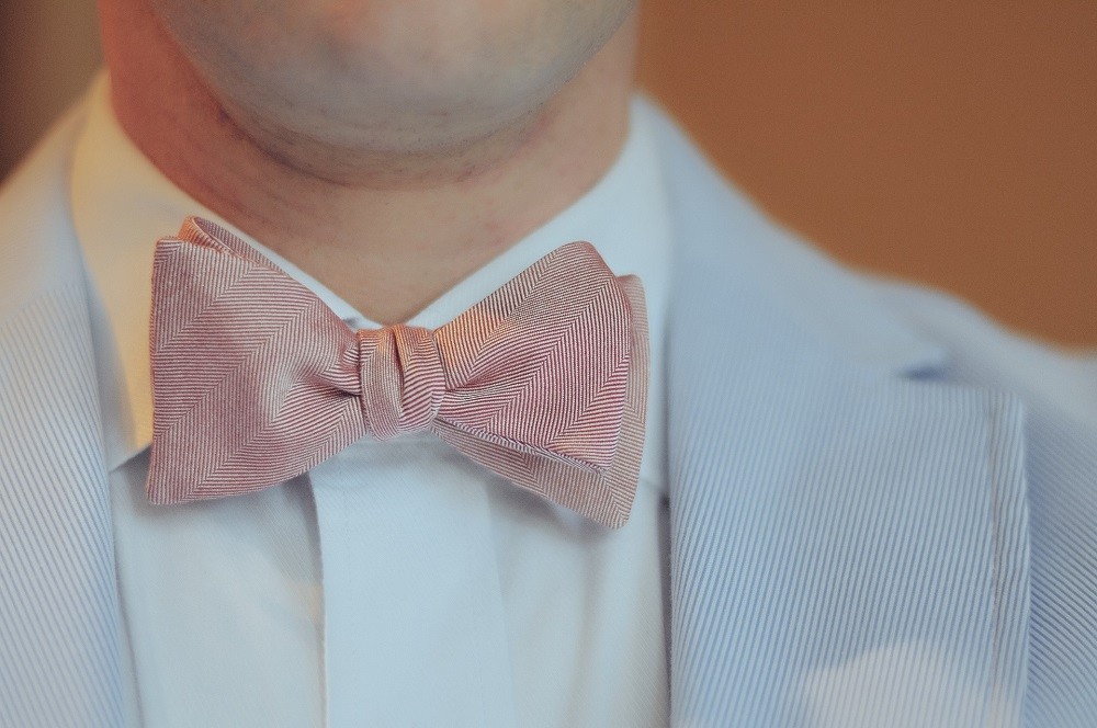A gentleman is wearing a red bow tie tied against a white collared shirt and a light blue suit jacket