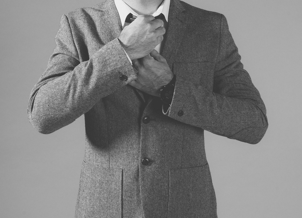 A black and white photograph of a man adjusting his tie. He is wearing a sport coat with all the buttons done up (this is seen as many as a fashion faux pas, as the bottom button of a sport coat should always be undone)