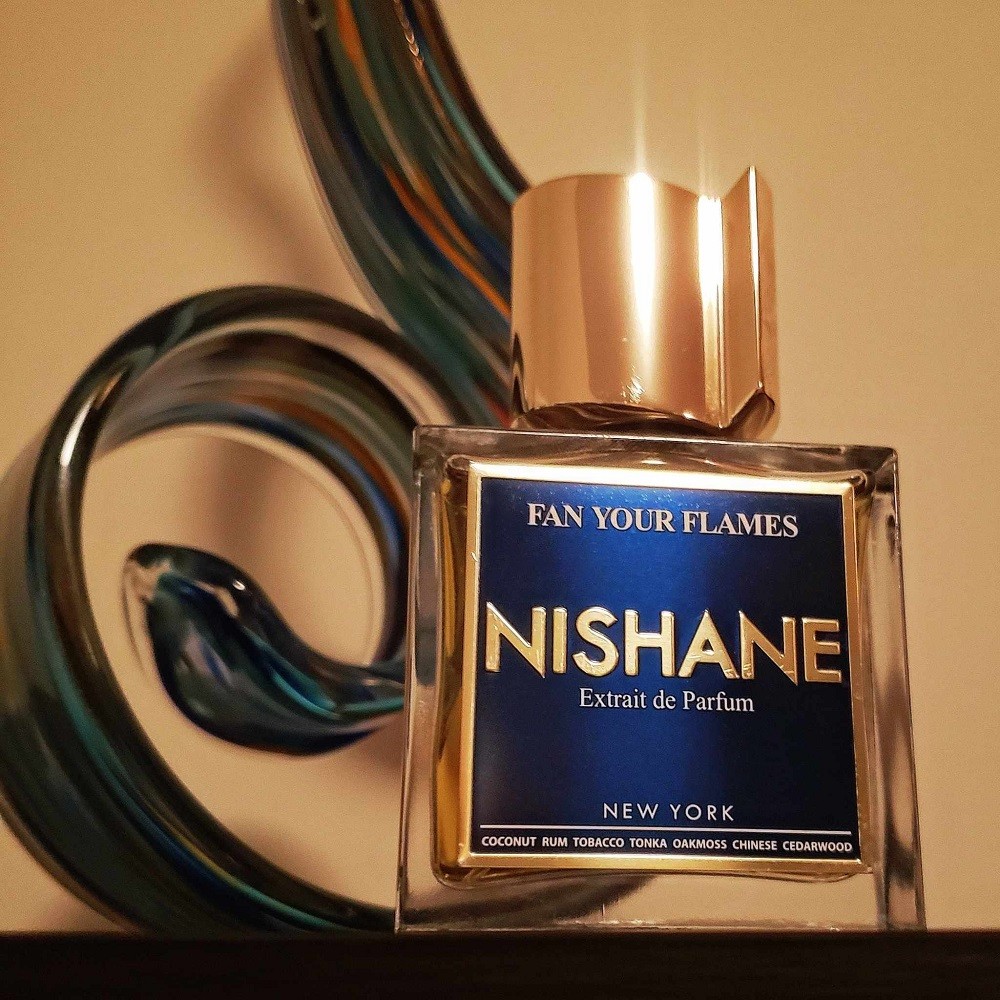 A square bottle of cologne with a large gold top sits in front of a blue blown glass flame. The bottle of cologne is Fan Your Flames by Nishane