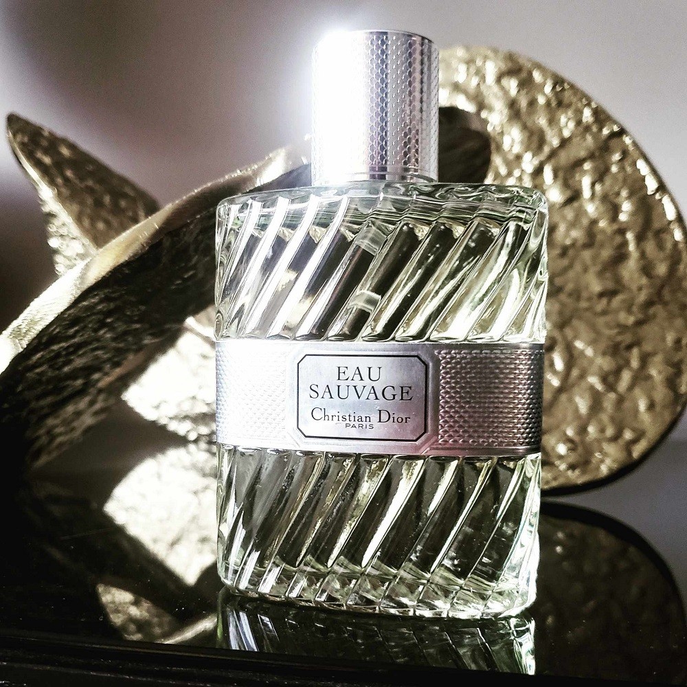Dior Eau Sauvage comes in a unique bottle with textured spirals that go vertically down the bottle. The name of the fragrance and brand is displayed on a silver band that spans horizontally across the middle of the bottle. The cap is silver, matching the band. The bottle sits on a glass table with a matte gold decoration in the background