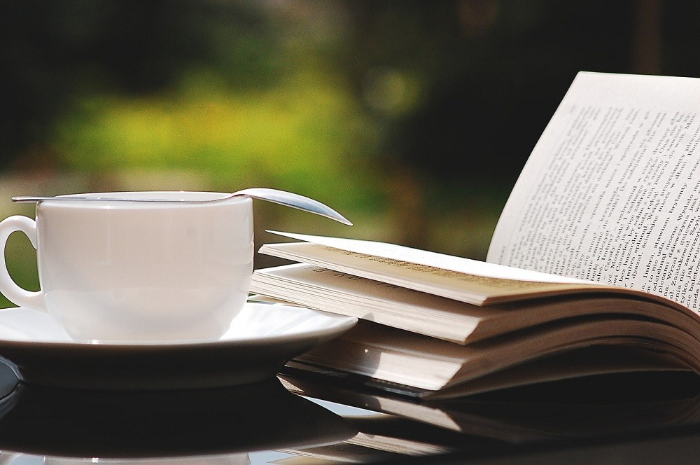 An open book sits beside a white coffee mug with a stirring spoon placed overtop. Both coffee mug and book are sitting on a table outside. There's a green shrub in the background and the sun is shining