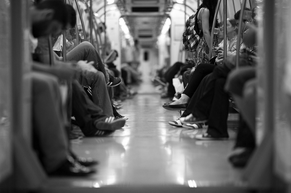 A black and white photograph showing many people riding a light rail train. The view is low so you only see their feet and part of their torsos