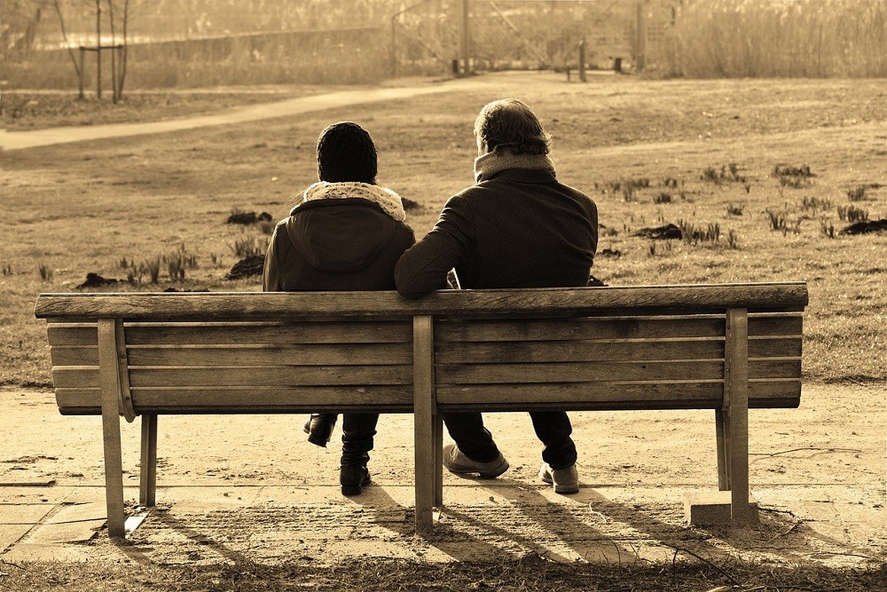A couple of friends facing away from the camera are sitting on a bench. They appear to be deep in conversation