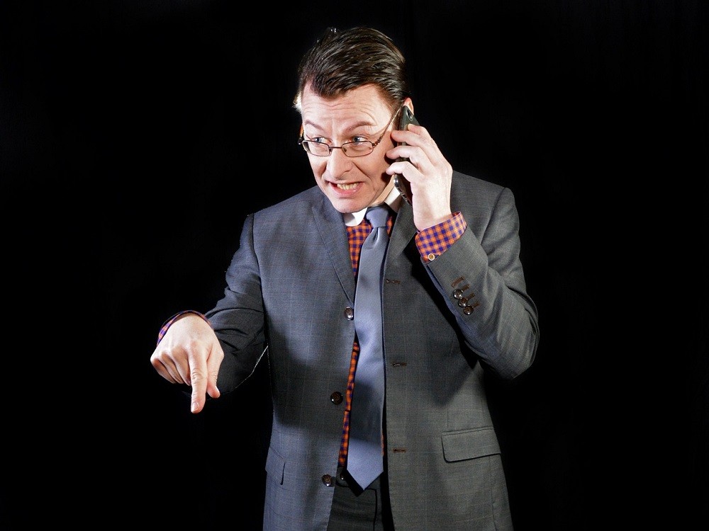 A middle-aged man wearing a suit and tie is yelling at someone on a cell phone. His face is scrunched up like he is in the middle of screaming at whoever is on the other line. His body language is very tense, including pointing down with his other hand