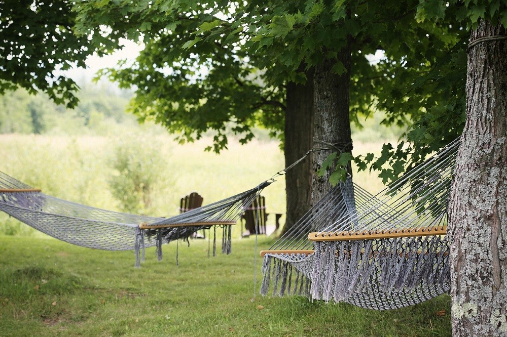 A string hammock is tied to a large tree. A person is sitting in it relaxing staring up at the clouds