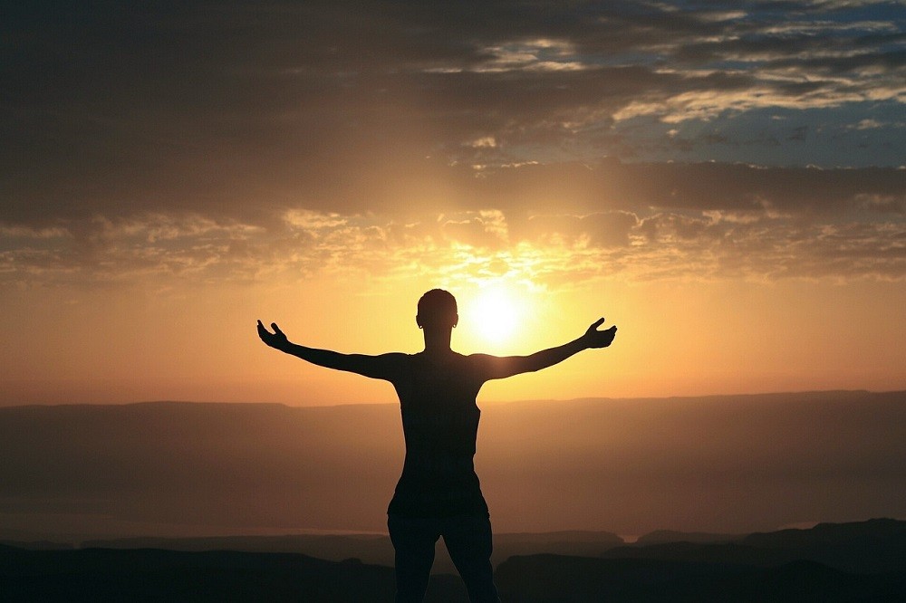 The sun is setting. A man with outstretched arms stands facing away from the camera, embracing the sun and warmth