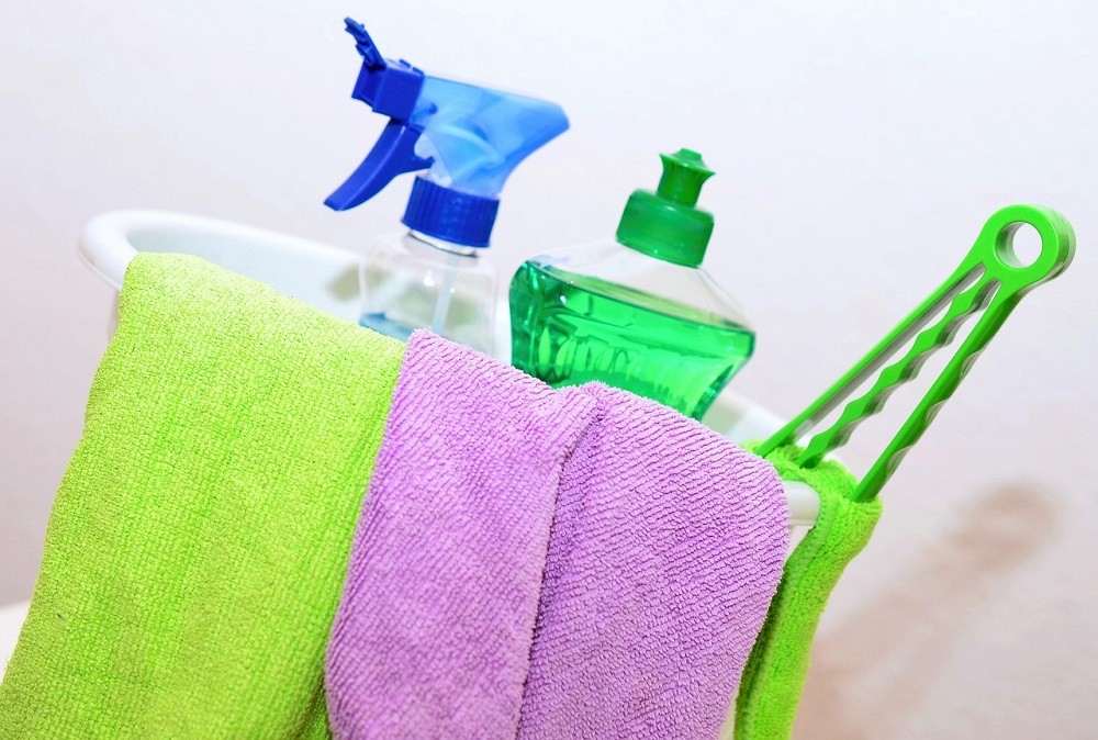 Various cleaning supplies including a bottle of dish soap and a spray bottle are in a bucket with two cloths draped over the edge of the bucket