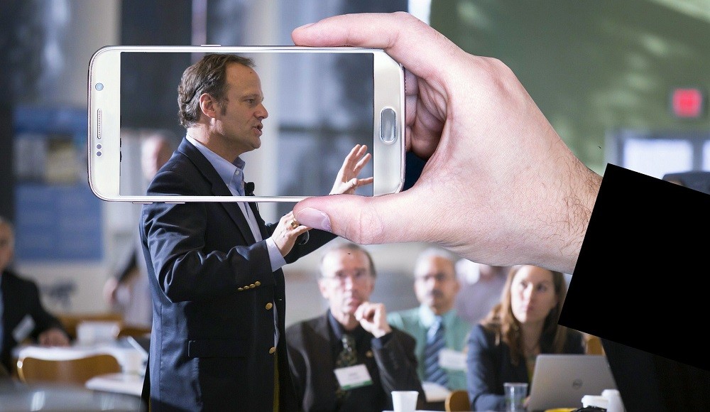 A smart phone is held sideways and whoever is holding it up is taking a video of a businessman speaking