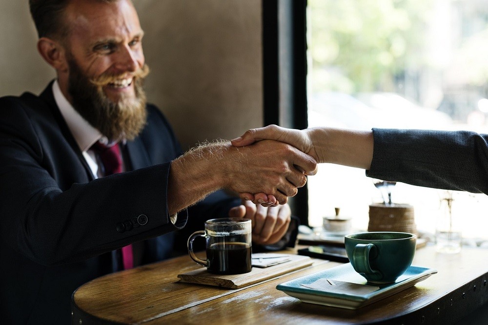 A man with a beard and moustache is wearing a suit. He's sitting at a coffee table and shaking hands with someone who is out of frame