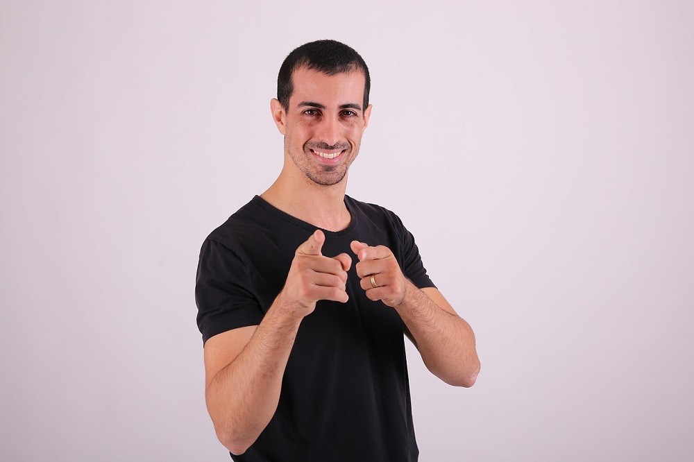 A middle aged man wearing a black t-shirt is smiling and pointing his fingers at the camera. He looks very happy