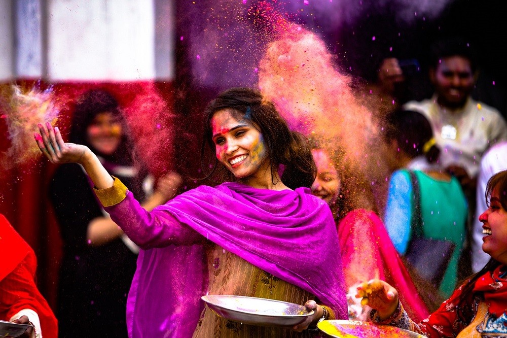 An Indian woman wearing a purple robe is throwing coloured powder in the air. It looks like she is celebrating with other people who are also throwing coloured powder