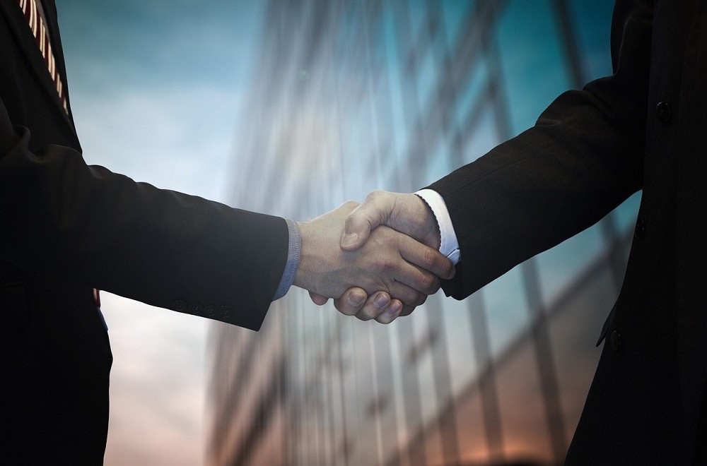 Two businessmen are shaking hands. Both are wearing black suits with white dress shirts poking out from the cuff of the arm hole Their torsos are out of frame. Their arms are outstretched and the handshake is professional