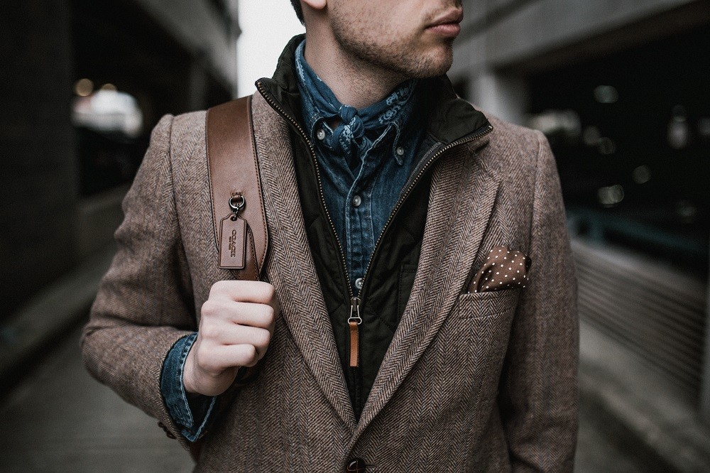 A younger man is wearing a light brown sport coat with a brown and white polka-dot pocket square and blue scarf. He looks very happy