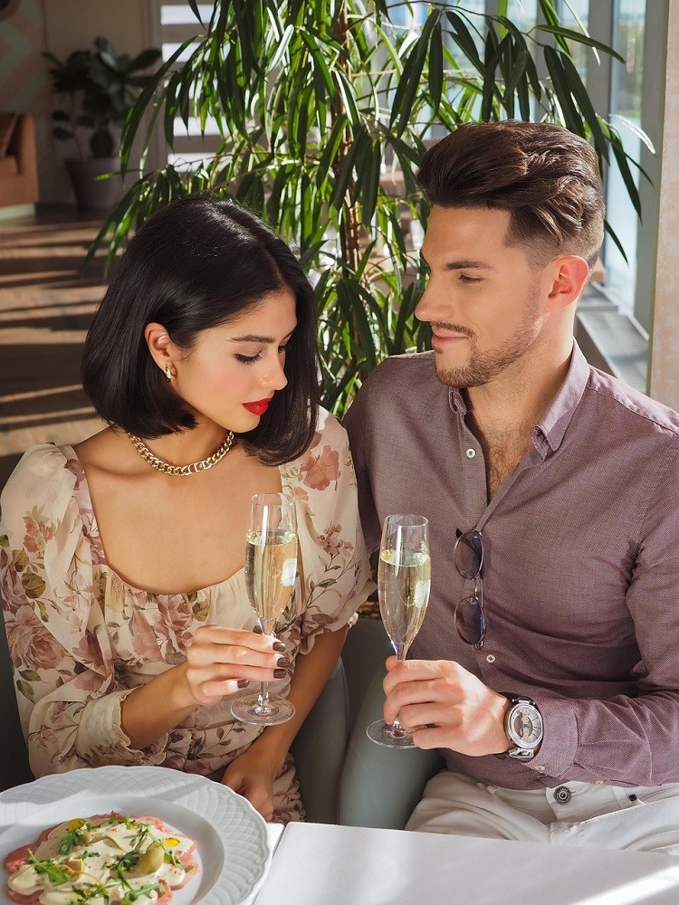 A young couple sit at a table at a restaurant. They have champagne flutes in their hands. The woman is wearing a floral summer dress and the man is wearing a purple dress shirt