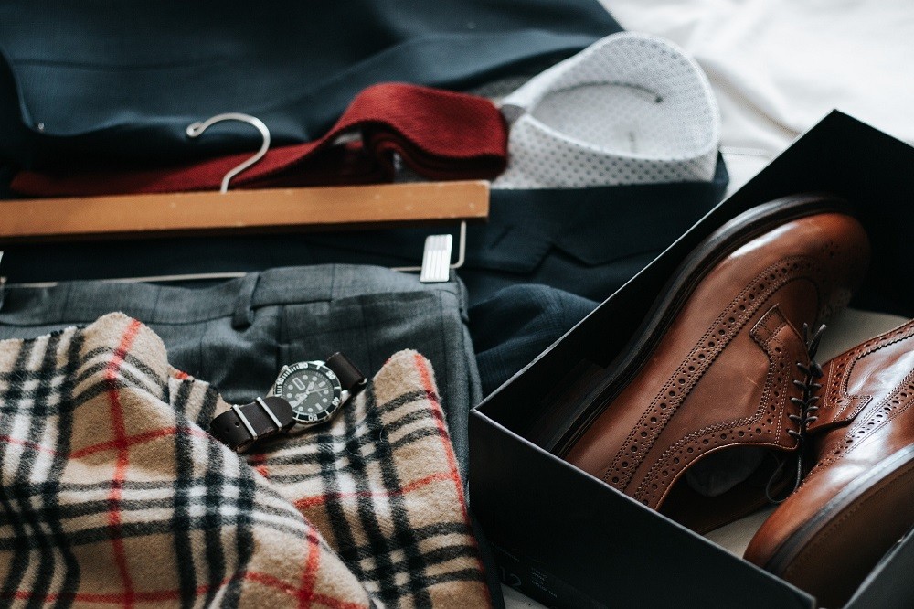 A pair of dark brown dress shoes, a fancy black dress watch, and dress clothes are displayed on a table