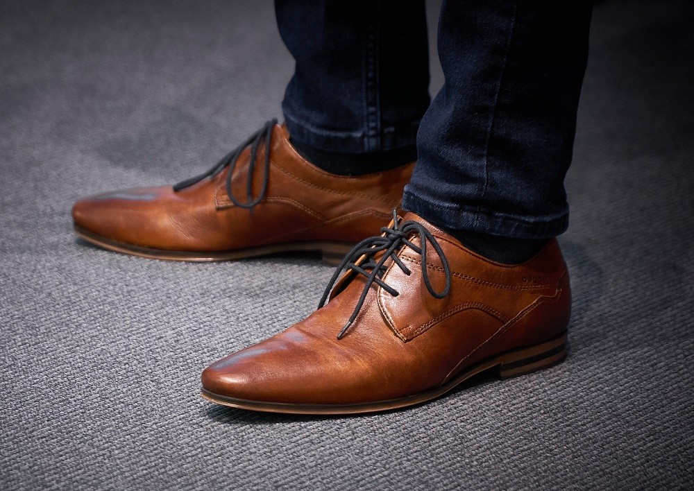 A man is wearing medium brown dress shoes and dark wash jeans. Only his shoes and the bottom part of his jeans are visible in the picture