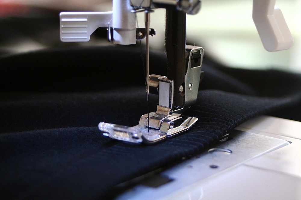 A sewing machine has a piece of fabric in it. The fabric appears to be dark blue with a subtle white pinstripe, perhaps a pair of trousers