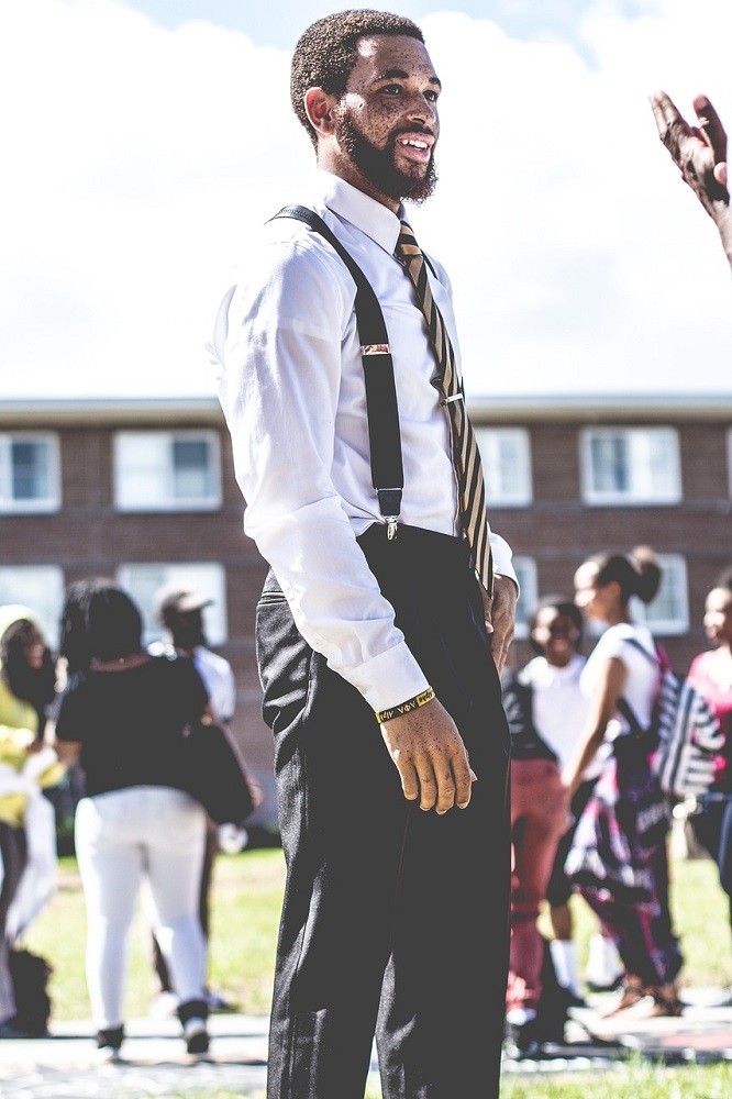 A younger Afro-American man is wearing a white dress shirt, black dress pants, and black suspenders. He is walking towards someone who is out of the frame