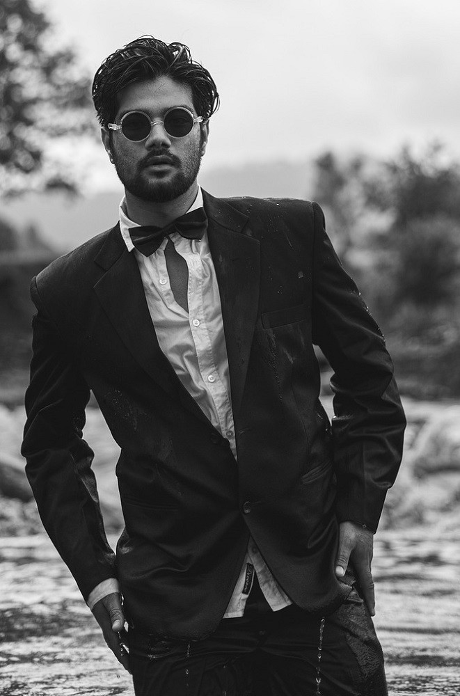 A younger man looks unkept. There are many style mistakes in this picture: his hair is a mess, his shirt is buttoned up improperly, his suit jacket has rips and stains, his zipper on his pants is down, his bow tie isn't tied properly, and he is trying to pose in a very unflattering manner with his hands on his hips