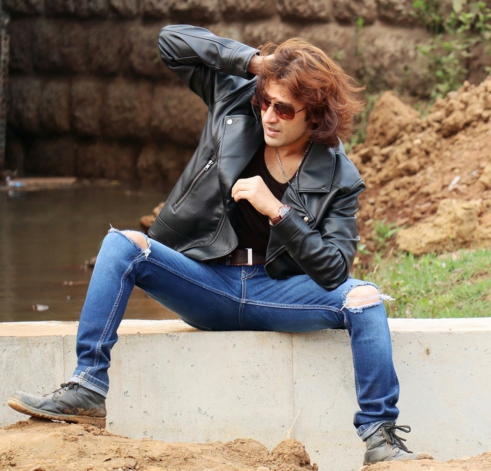A younger man with longer brown hair is posing wearing a leather jacket and ripped blue jeans