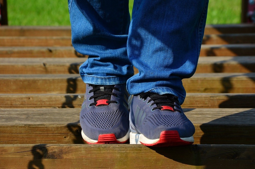 A pair of bright blue and red running shoes are worn. The person wearing these shoes is also wearing jeans. They are walking across a wooden bridge or boardwalk