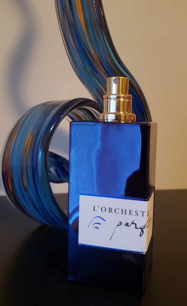 A rectangular bottle of cologne. It has a shiny blue colour to it and a gold atomizer. A white label is on the front. It has the company name along with the name of the fragrance