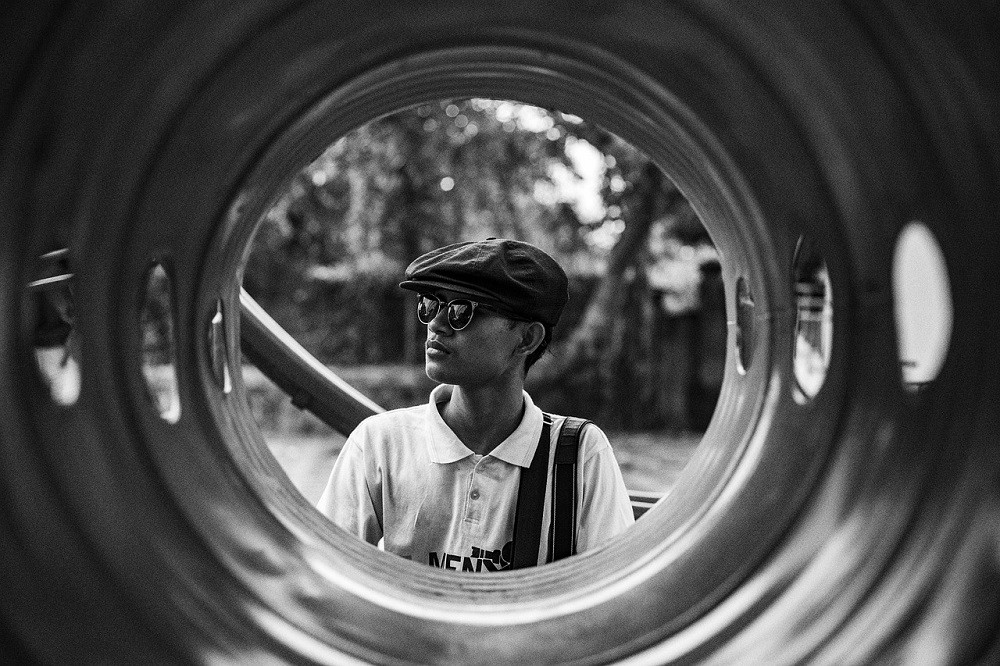 A black and white photograph of a younger man wearing a flatcap and sunglasses. He is positioned at the end of some sort of industrial tubing with the photographer on the other end, creating a unique viewpoint where the man is the focal point and the tube is a border around him