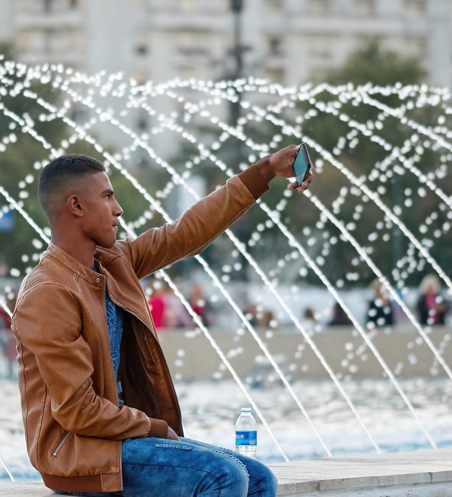 A young man wearing a brown leather jacket and blue shirt is sitting in front of a fountain. He is taking a selfie
