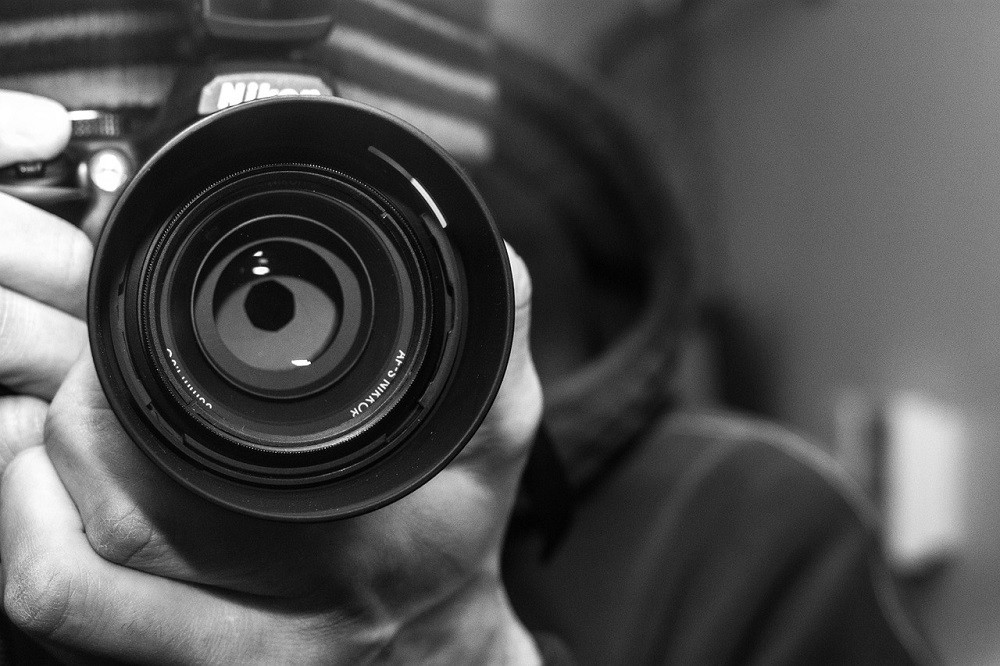 A black and white photograph of a person holding a professional Nikon brand camera up as if they are about to take a picture