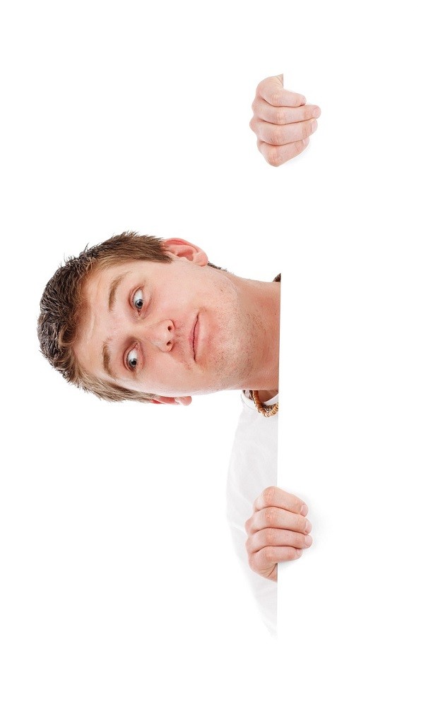 A man making a silly face is peeking out from behind a white wall