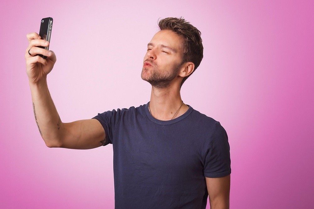 A younger man is taking a silly selfie. He's wearing a navy blue tee shirt and making a silly duck face at his phone