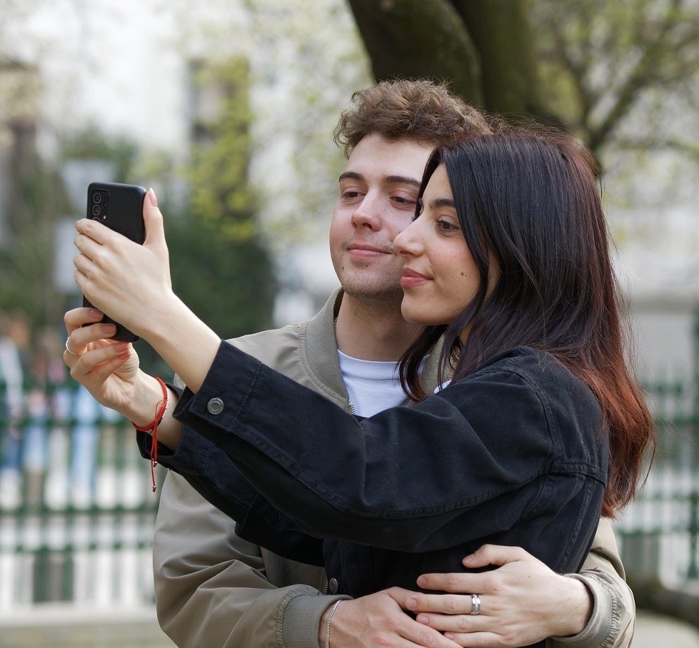 A young couple poses for a cute selfie. She has long brown hair and is wearing a black hoodie. He has short curly brown hair and is wearing a tan shirt