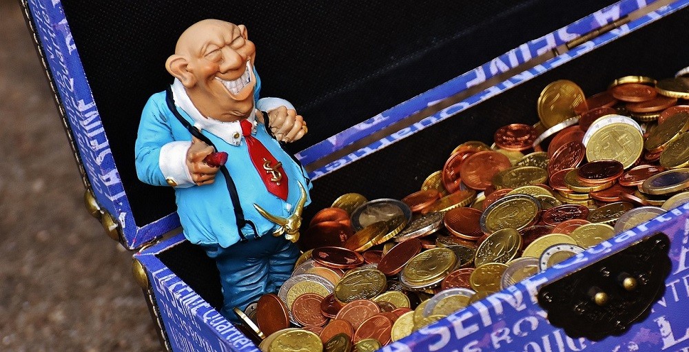 A small figurine of an old, heavy-set man with a dollar sign on his tie and a cigar in his hand is positioned inside a trunk full of coins, almost like he's guarding it