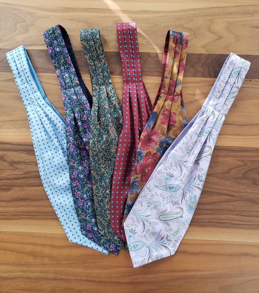 Multiple ascots are laid out on a table. Some have floral patterns, others have polka-dots, and some have paisley patterns. They are all brightly coloured (but not tacky or over-the-top)