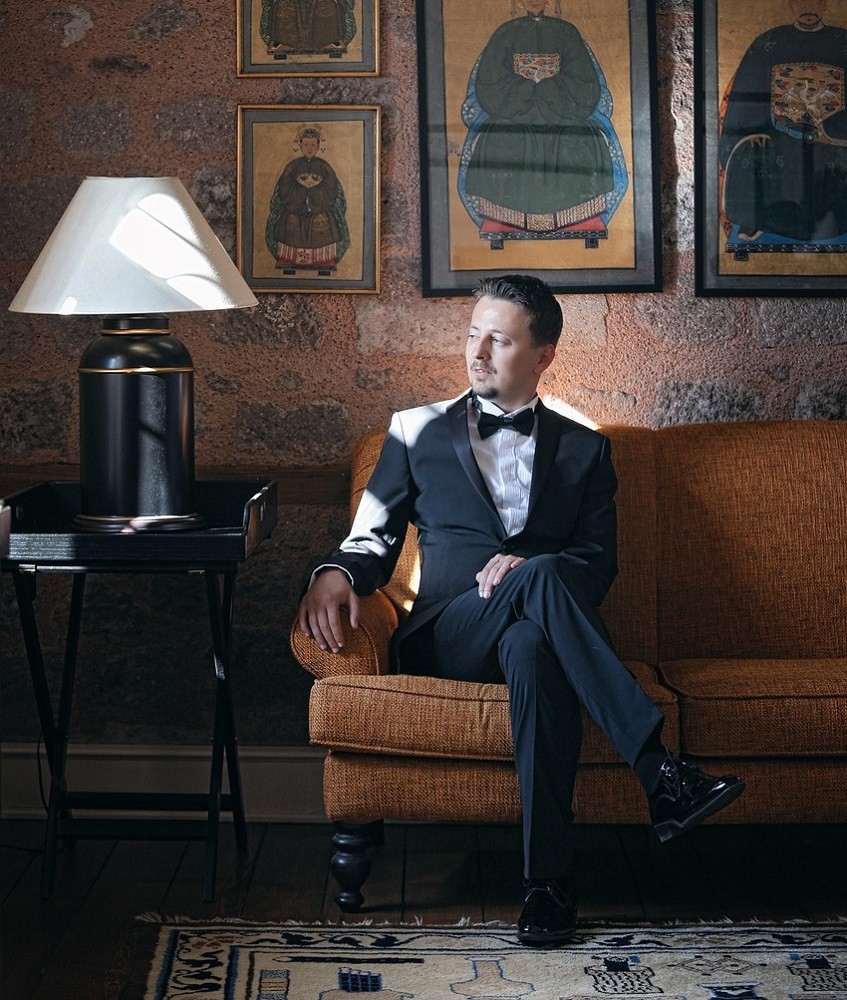 A gentleman in a suit sits on a large couch. He is dressed in black tie attire, complete with a black bow tie