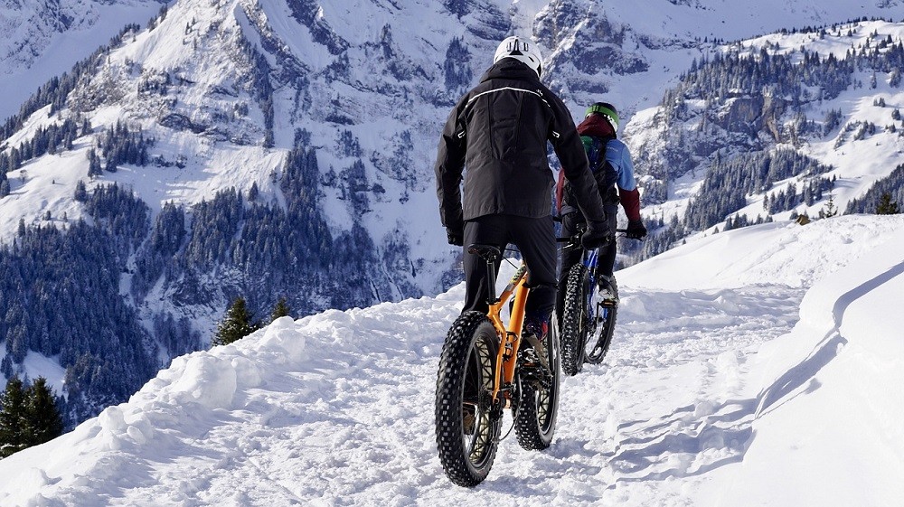 A couple of bikers (possibly on a date) are riding along some snowy terrain. They are both wearing full winter weather gear and their bikes have large tires on them