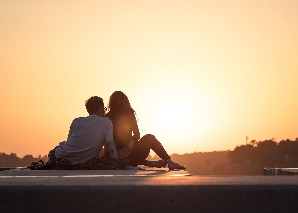 A young couple sits close together watching the sun set. The man has his arm around the woman