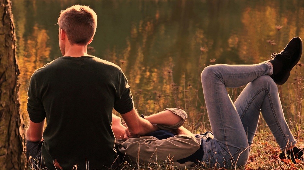A young couple are on a date sitting beside a tree. The woman is laying down with her head on the mans leg. He has his back against the tree and legs out straight