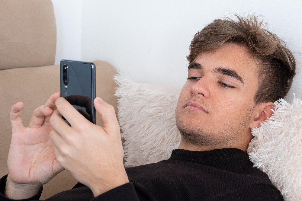 A young man is laying down on a couch. He is checking his text messages on his phone