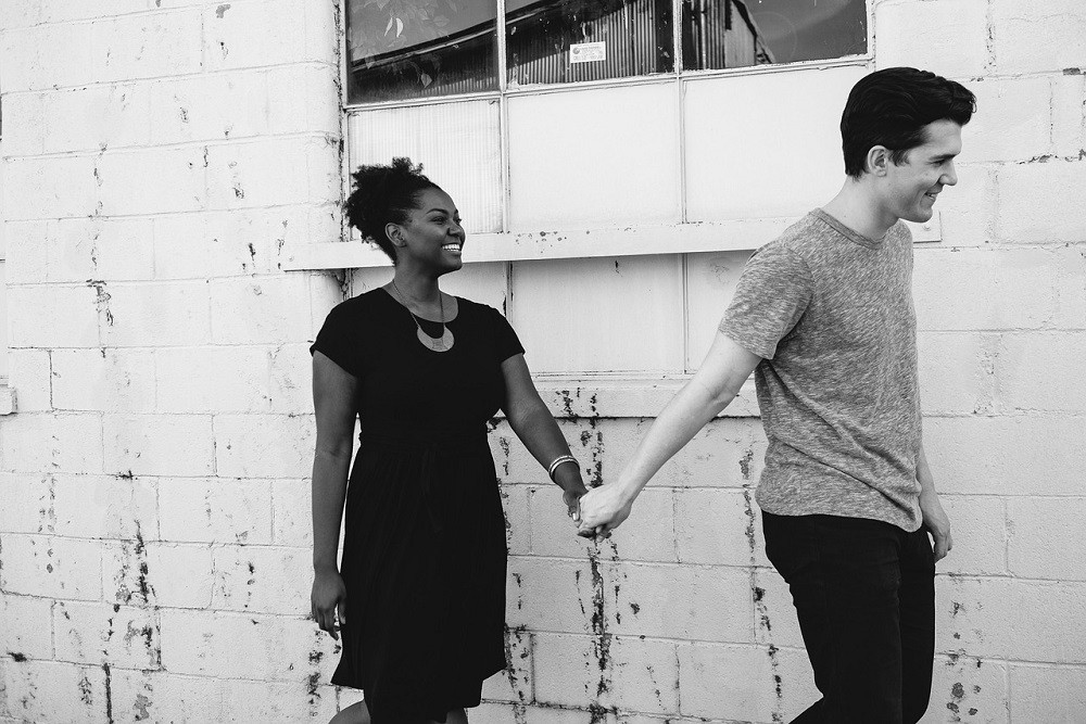 A young couple hold each others hand. The man is walking in front of the woman on a narrow sidewalk