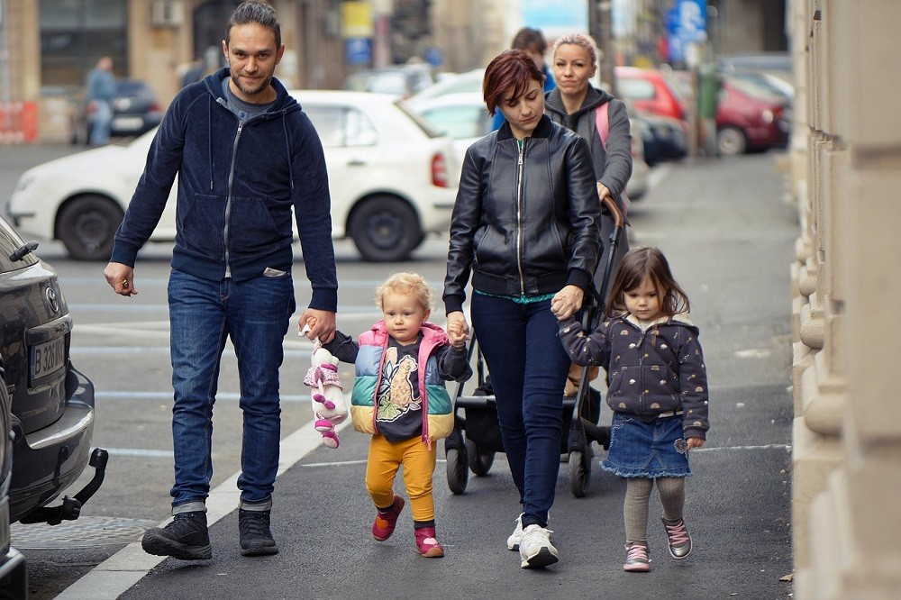 A family consisting of a man, woman, and two young daughters are walking on a sidewalk. The husband is walking closest to the road