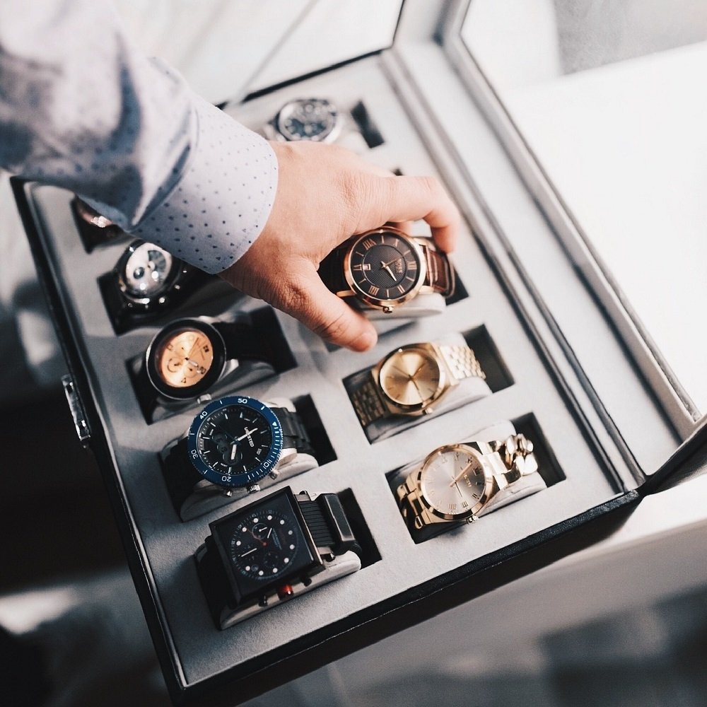 A man is placing a watch into a watch box. The watch itself is wrapped around a cushion that will be slid into a slot inside the watch box, protecting it from damage